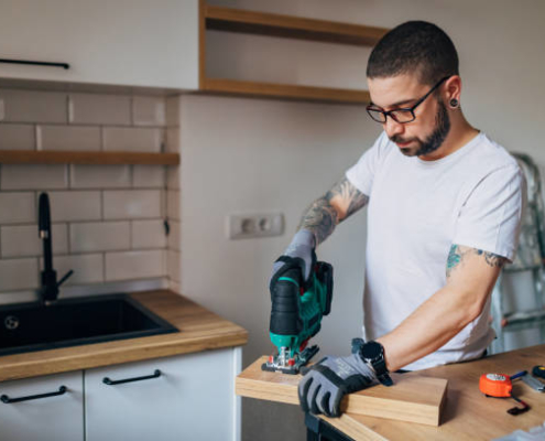 Skillful carpenter in white T-shirt using jigsaw to renovate the kitchen.